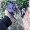 A person holding a Large Amethyst Dragon’s Tooth Scepter from Bahia, Brazil 902 grams in front of a brick wall.