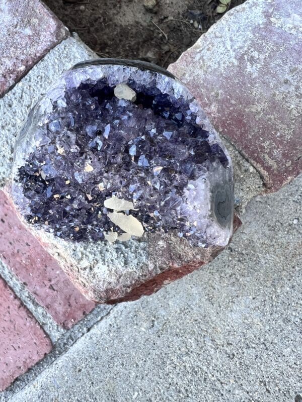 An Uruguayan Amethyst Cut Base with Secondary Calcite 822 grams sitting on a brick sidewalk.